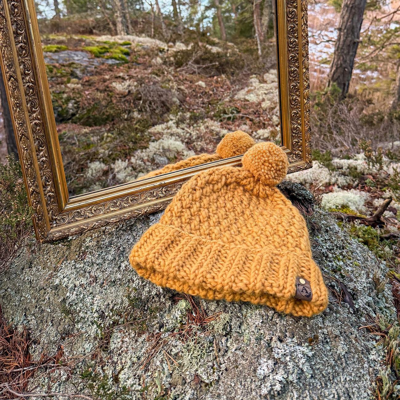 Mustard yellow beanie with twin pompoms and a leather tag, on a lichen covered rock in front of the forest mirror