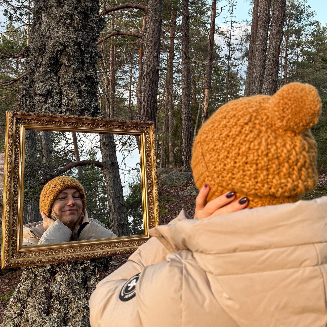 Wearing the yellow beanie, looking into the mirror hung on a pine