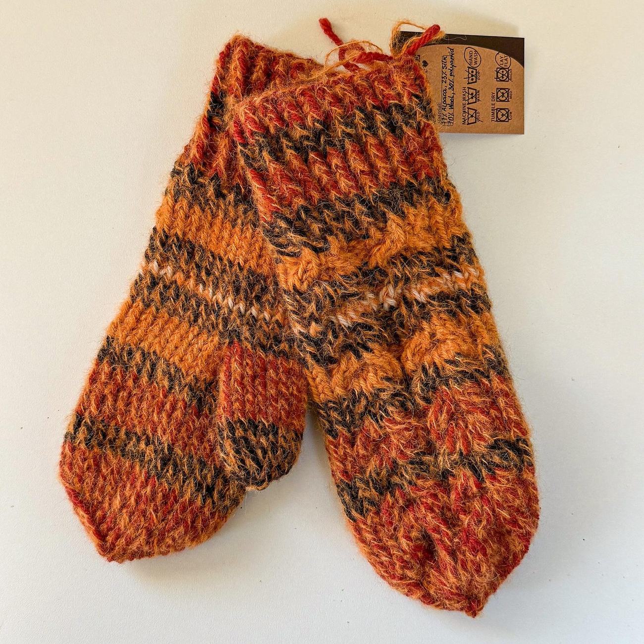 Wool and alpaca mittens in orange, rust and charcoal with a white stripe, laid flat with the care tag
