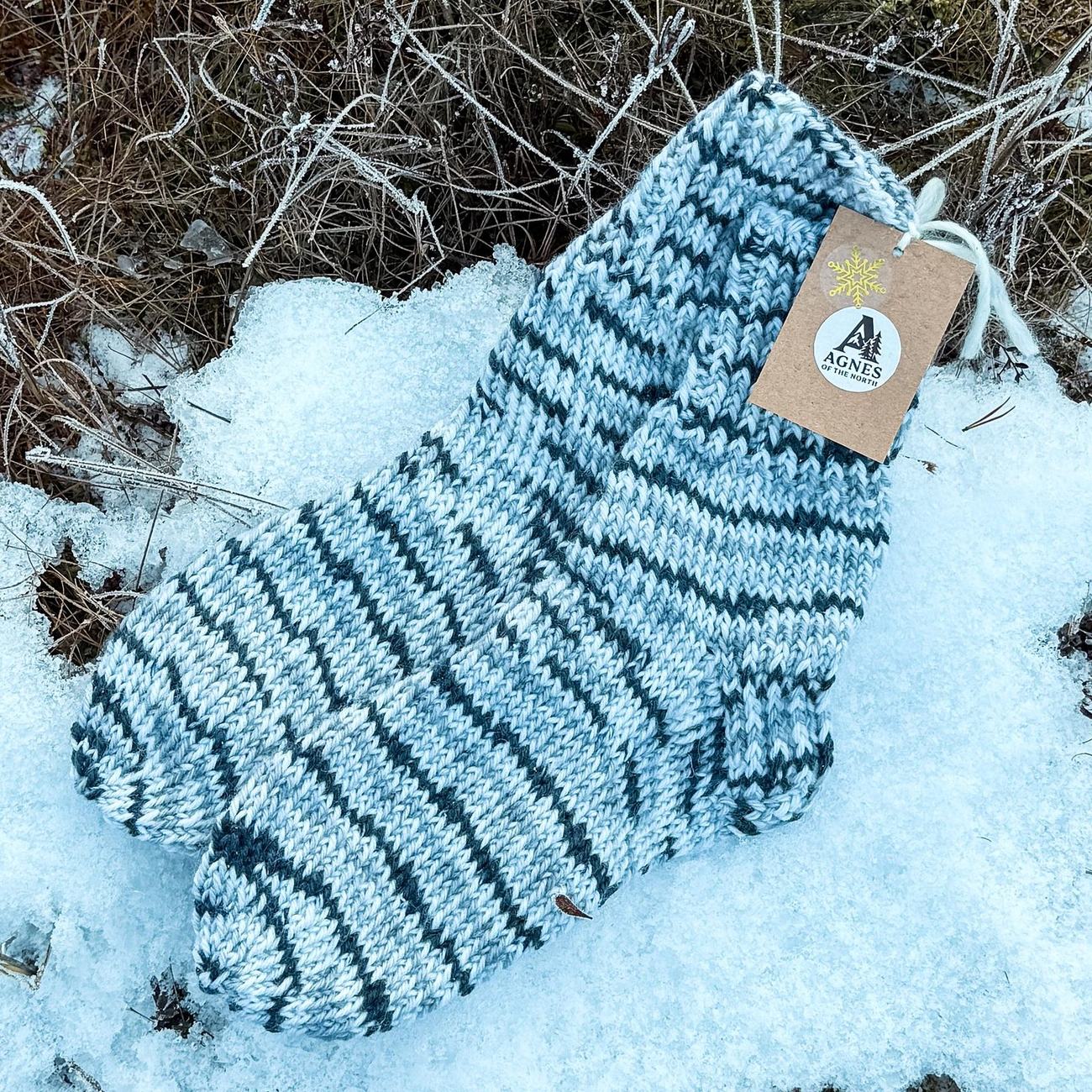 Grey melange socks with charcoal stripes and the Agnes of the North tag, lying on fresh snow