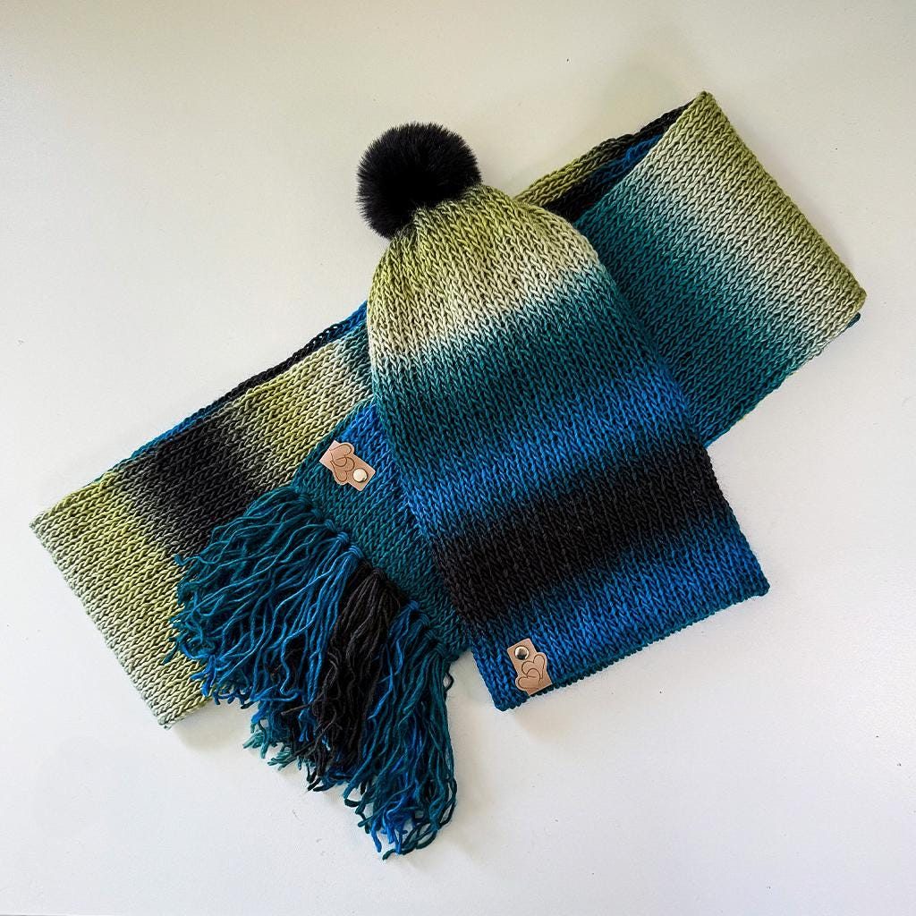 Beanie and scarf set in the Deep Forest gradient, olive green fading through teal and ocean blue to charcoal, with a black faux-fur pom and fringed ends