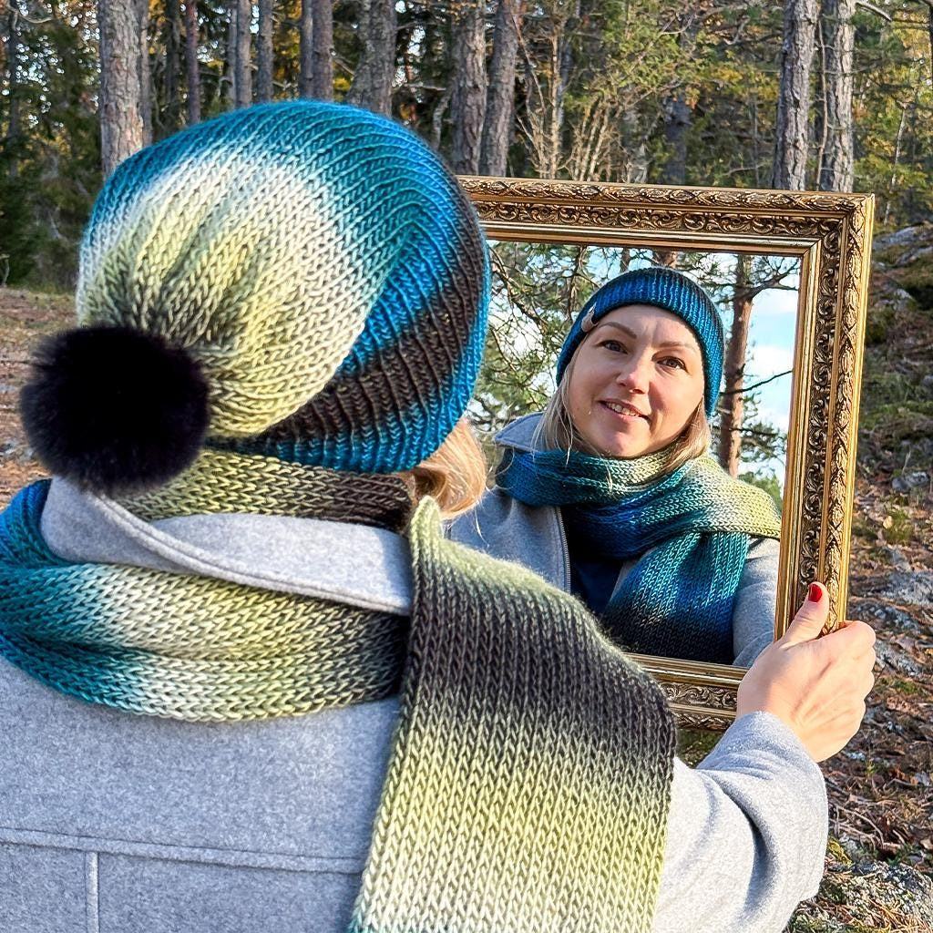 Agnes holding the gilded frame around herself, wearing the Deep Forest beanie and scarf among the pines